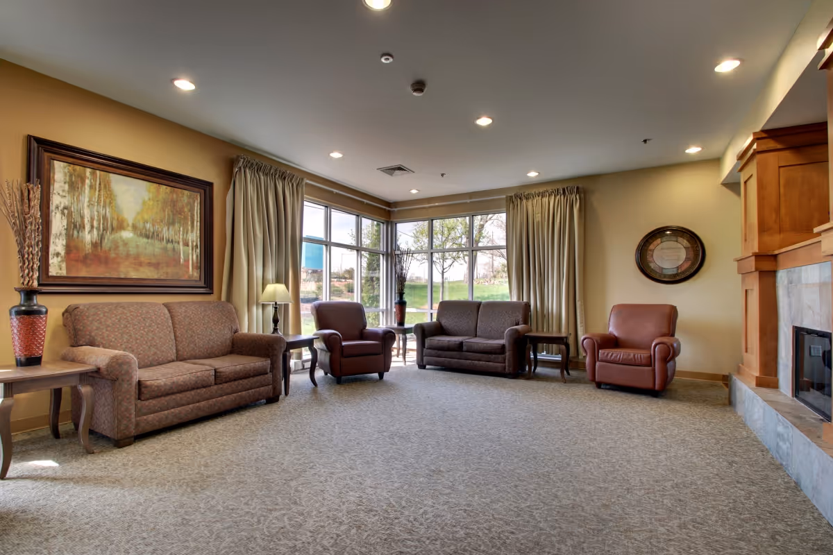 A spacious living room area with beige walls and carpeted floor. The room features two fabric sofas, two leather armchairs, wooden side tables with lamps, a large framed painting on the left wall, large windows with beige curtains letting in natural light, and a stone fireplace with a wooden mantle on the right.
