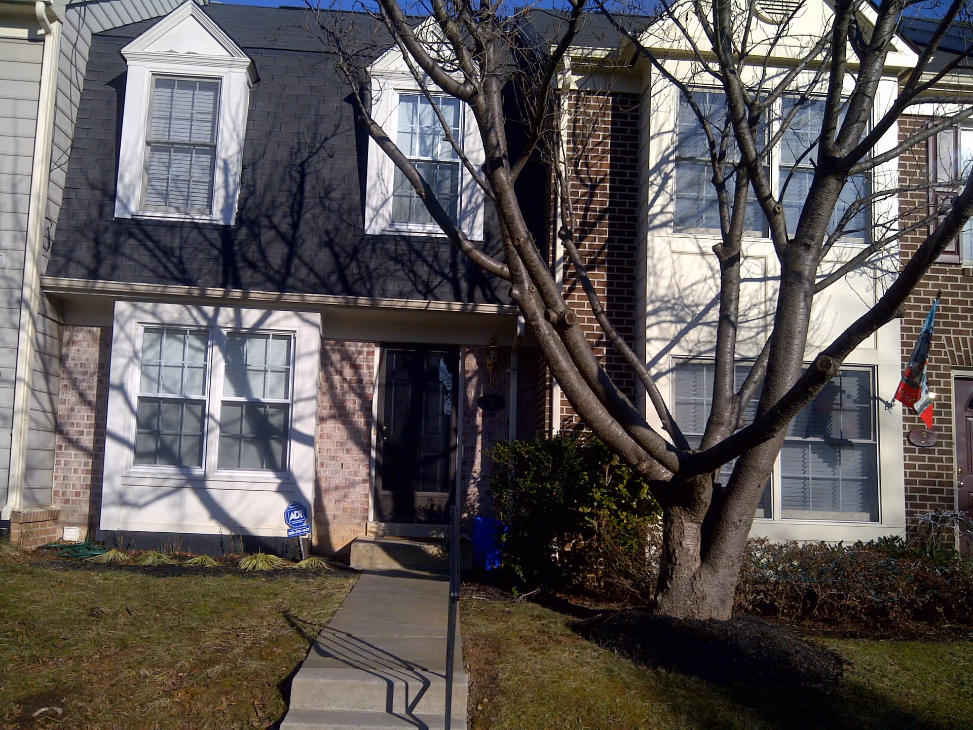 Front exterior view of a two-story brick townhouse with white window frames and a black front door. There is a large leafless tree in the front yard casting shadows on the building. A concrete walkway leads to the entrance, and there is a small garden area with some plants and a security sign near the front steps.