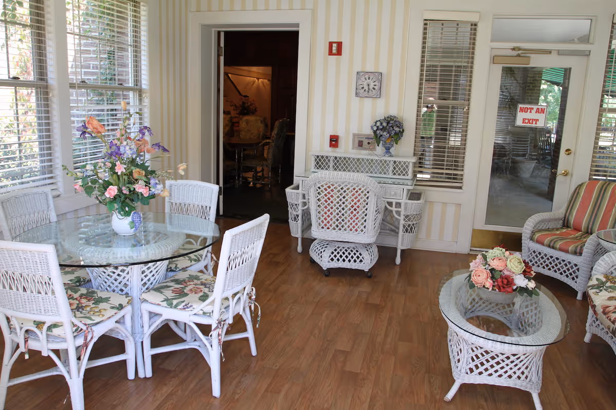 A bright and cozy sitting area with white wicker furniture including a round glass-top table with floral cushioned chairs, a wicker desk with a chair, and a glass-top coffee table with a floral arrangement. The room has large windows with blinds, striped wallpaper, and a door with a 'NOT AN EXIT' sign.