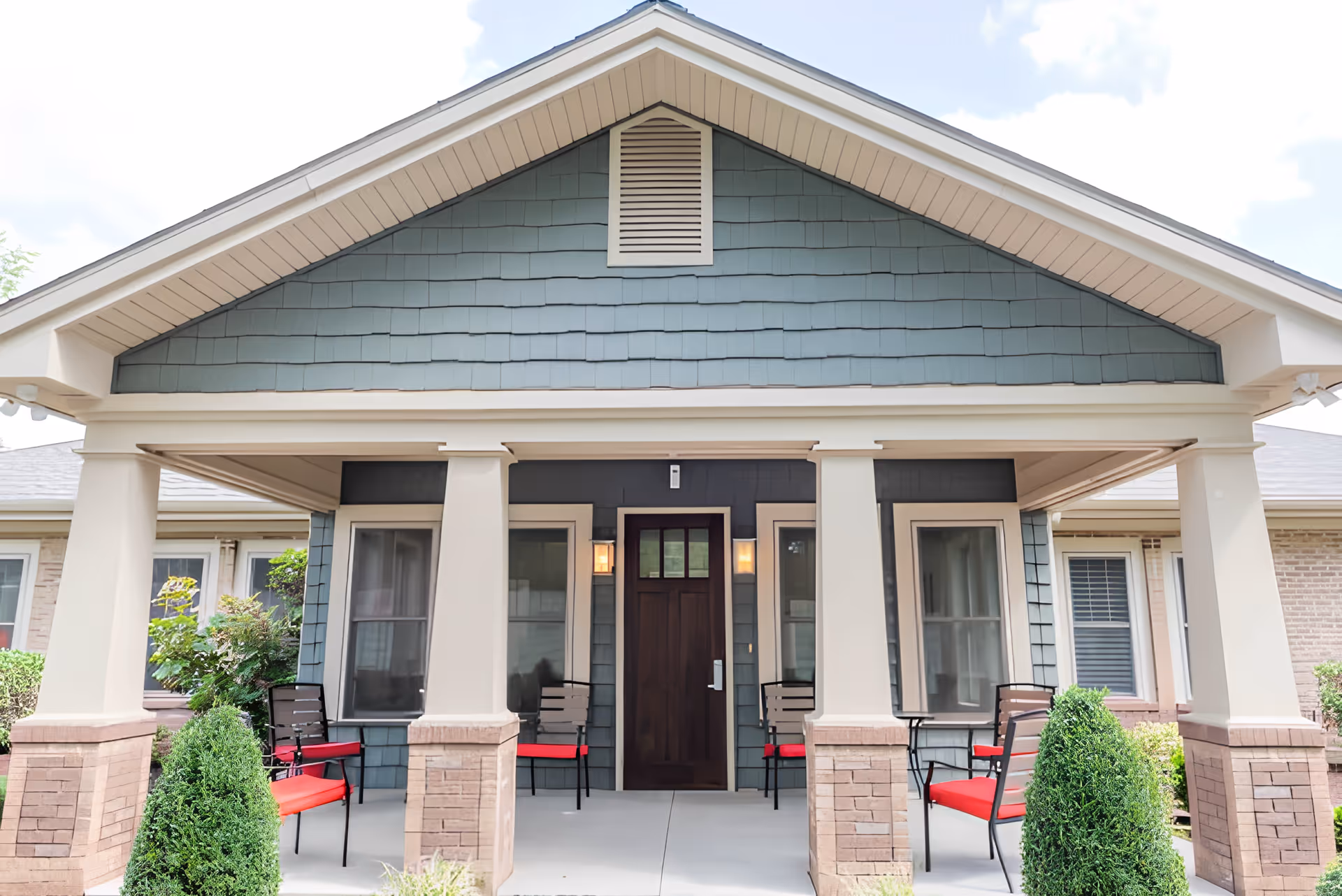 Front porch of a residential-style building with a dark wooden door centered between two windows. The porch has four beige columns with brick bases, and four black chairs with red cushions are arranged on the porch. There are neatly trimmed bushes in front of the porch and a blue-gray shingled gable above the entrance.