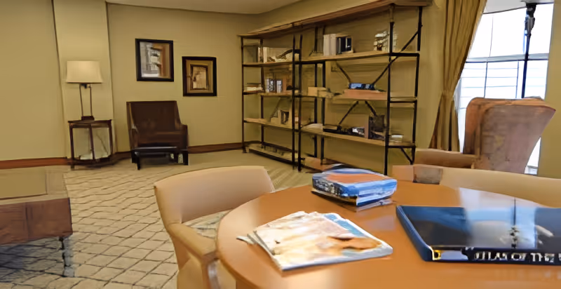 Cozy common room with a round table and chairs in the foreground, shelving with books and lounge chairs by a window.