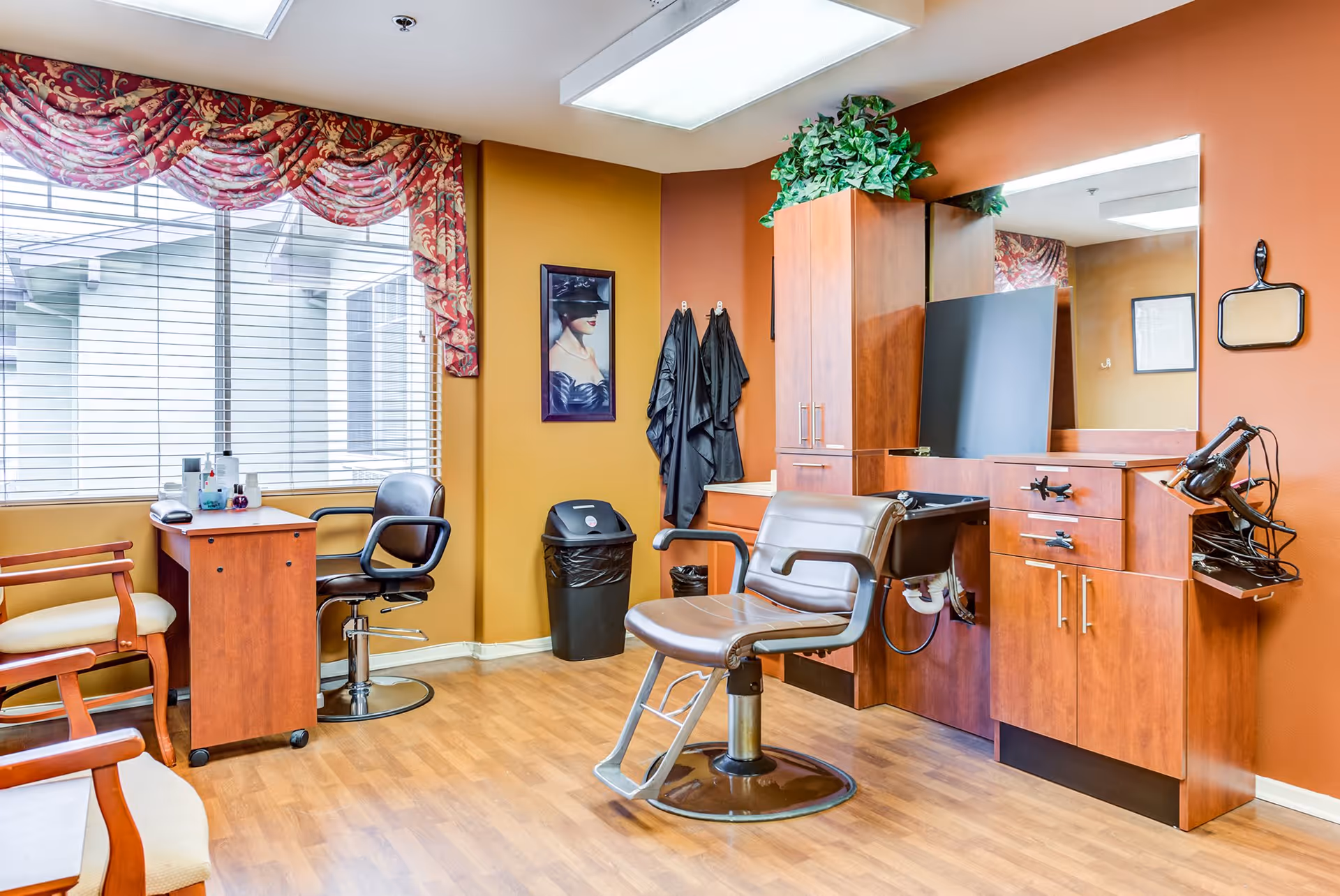 Interior of a salon room with a styling chair in front of a large mirror, wooden cabinets, a hair dryer, and a small desk with a chair near a window with red patterned valance curtains. The walls are painted in warm tones and there are two wooden chairs with cream cushions along one wall.