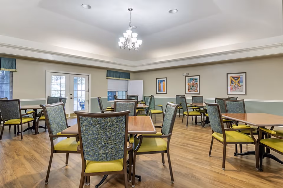 A dining room with multiple wooden tables and chairs featuring green and blue upholstery. The room has wooden flooring, beige walls with a green lower section, three framed artworks on the wall, a chandelier hanging from the ceiling, and double glass doors leading outside.
