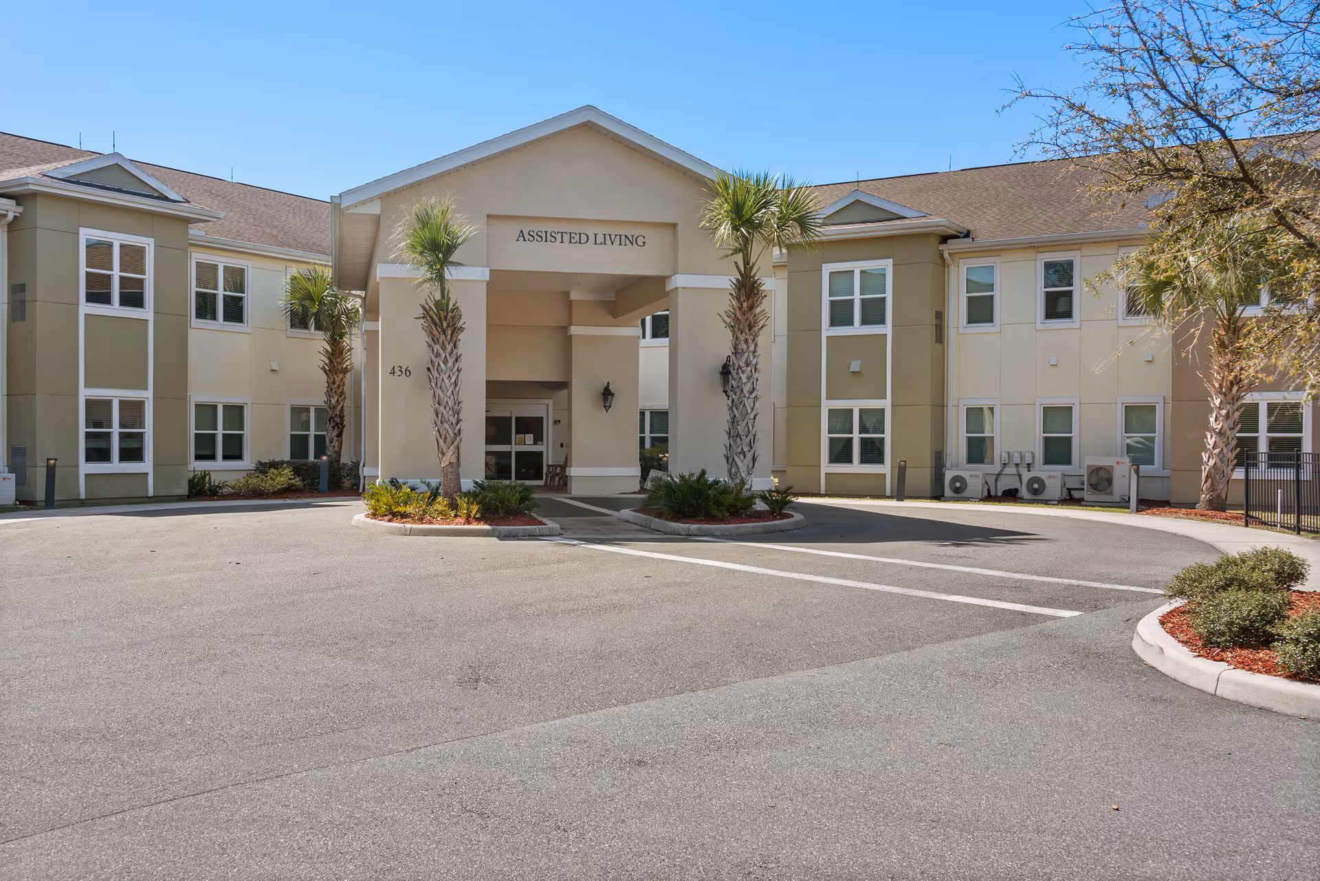 Front entrance of a two-story assisted living facility with a porte-cochère, palm trees, and a circular driveway.