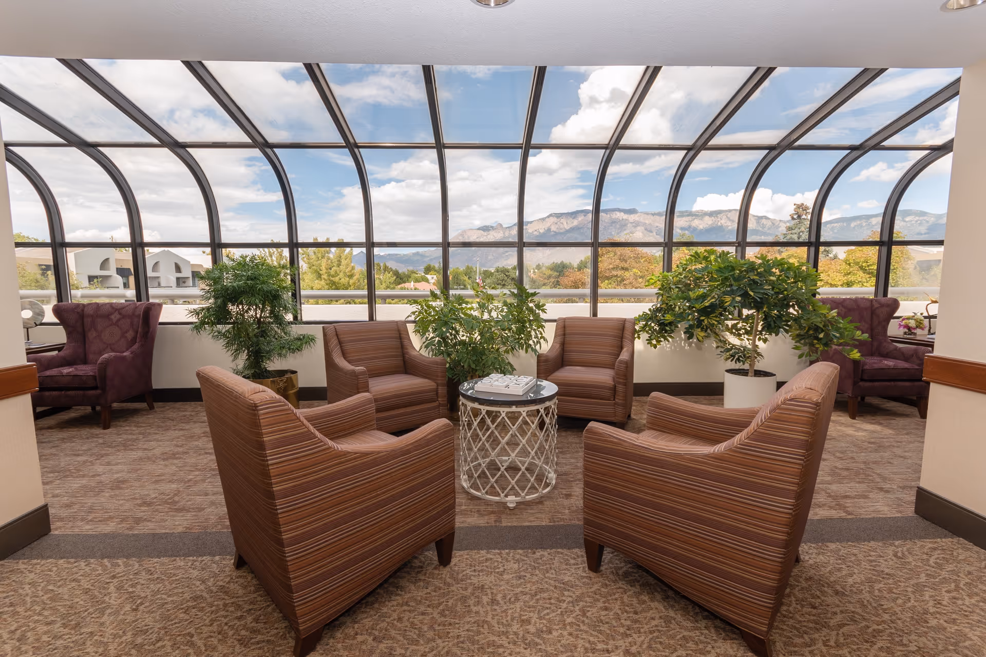 A bright indoor seating area with a curved glass ceiling and large windows offering a view of mountains and trees outside. The space features four striped armchairs arranged around a small round table with a decorative item on top. There are also two purple armchairs and several potted green plants placed near the windows.