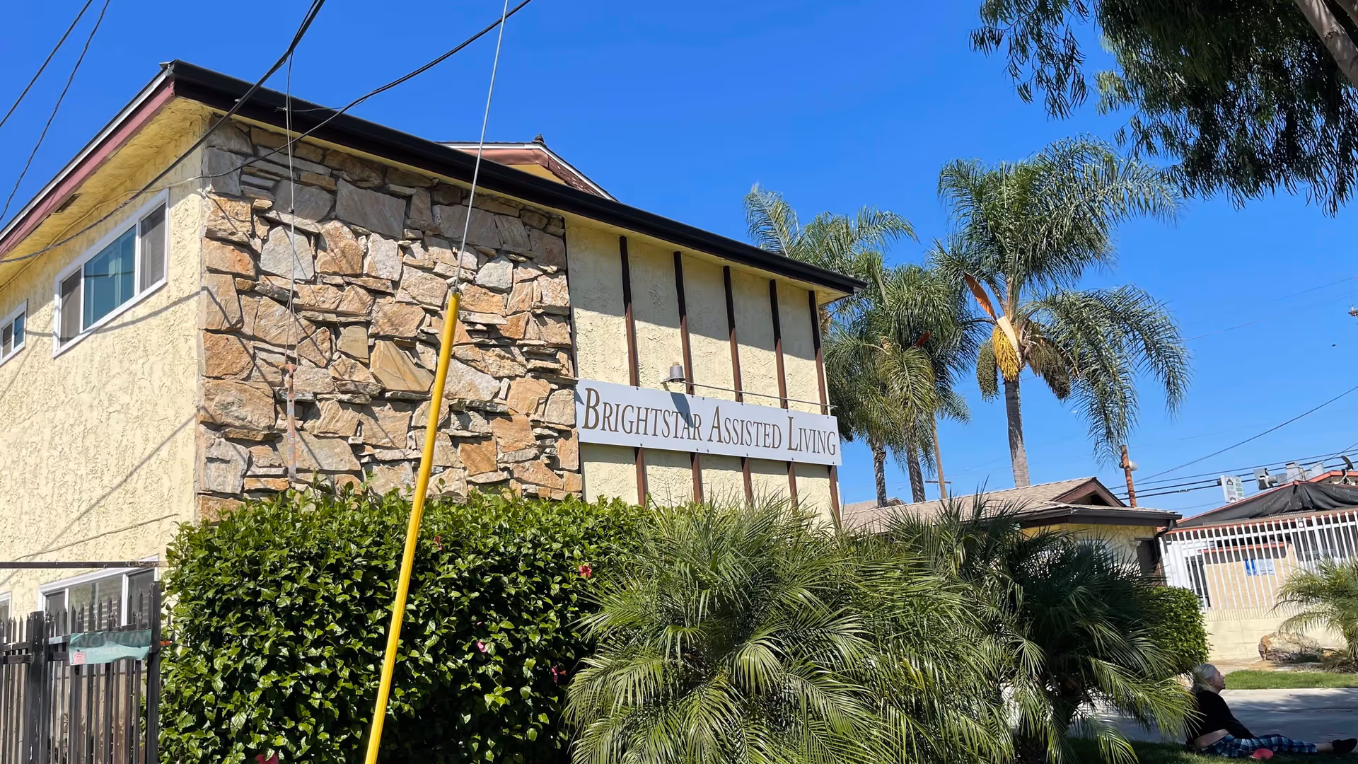 Exterior view of a two-story assisted living facility building with a stone and stucco facade, a sign that reads 'Brightstar Assisted Living,' surrounded by green bushes and palm trees under a clear blue sky.