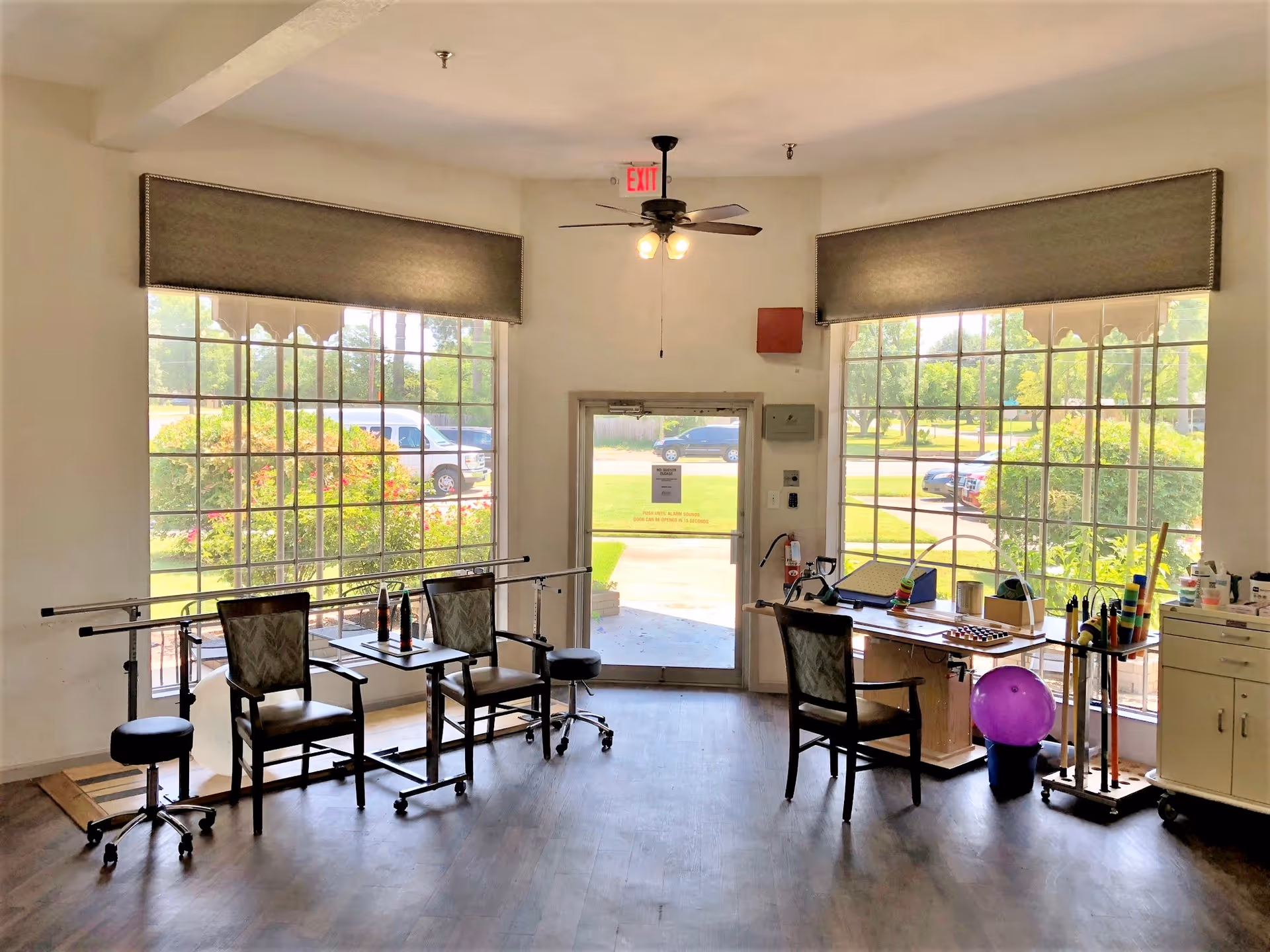 A bright room with large windows showing a green outdoor area. Inside, there are chairs, a table with various therapy or activity items, a purple exercise ball, and handrails along the wall, suggesting a physical therapy or rehabilitation space.