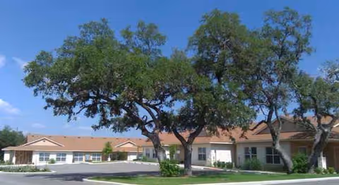 One-story tan senior living building with multiple entrances and large trees in front.