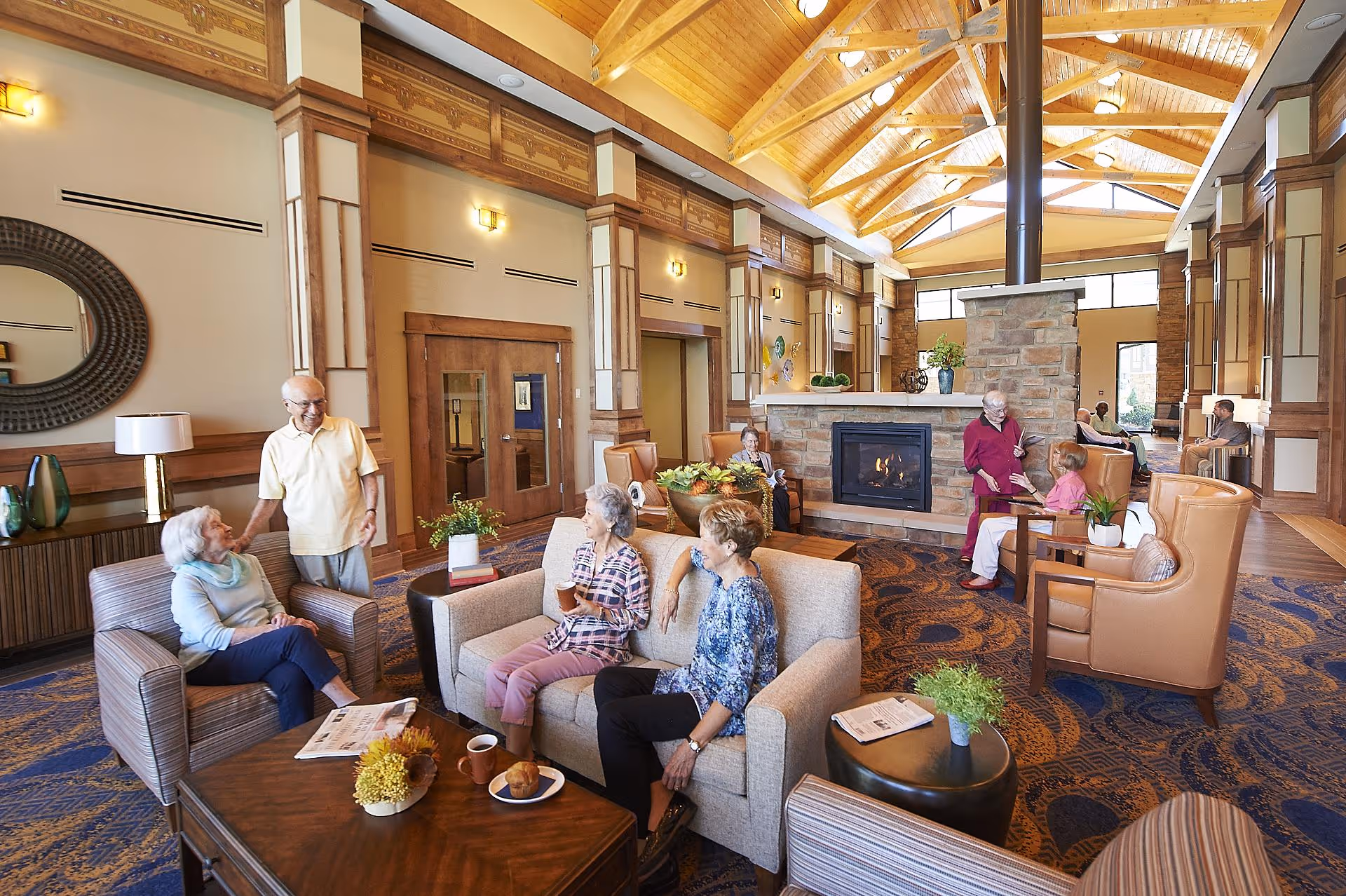 Seniors socializing in a spacious, warmly lit lounge with a stone fireplace and a high wooden vaulted ceiling.