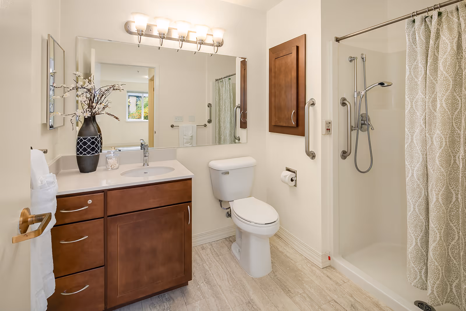 A clean and well-lit bathroom featuring a wooden vanity with a white countertop and sink, a large mirror above the sink with a row of lights, a white toilet, and a walk-in shower with a patterned shower curtain. There is a decorative vase with branches on the countertop and a towel hanging on a rack beside the vanity.