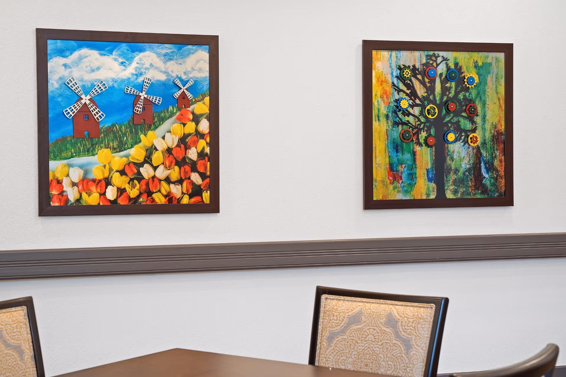 Two colorful framed paintings hanging on a white wall above a wooden table with patterned chairs. One painting depicts three windmills with a field of tulips under a blue sky with clouds. The other painting shows a tree with colorful gears as leaves on a multicolored abstract background.