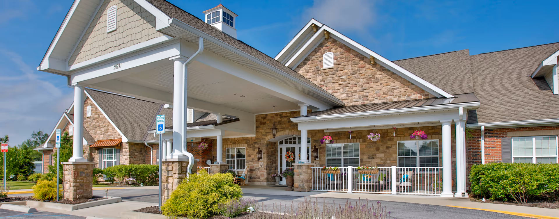 Front exterior view of Bickford of Greenwood facility showing a covered entrance with white columns, stone and brick facade, hanging flower baskets, and a clear blue sky.