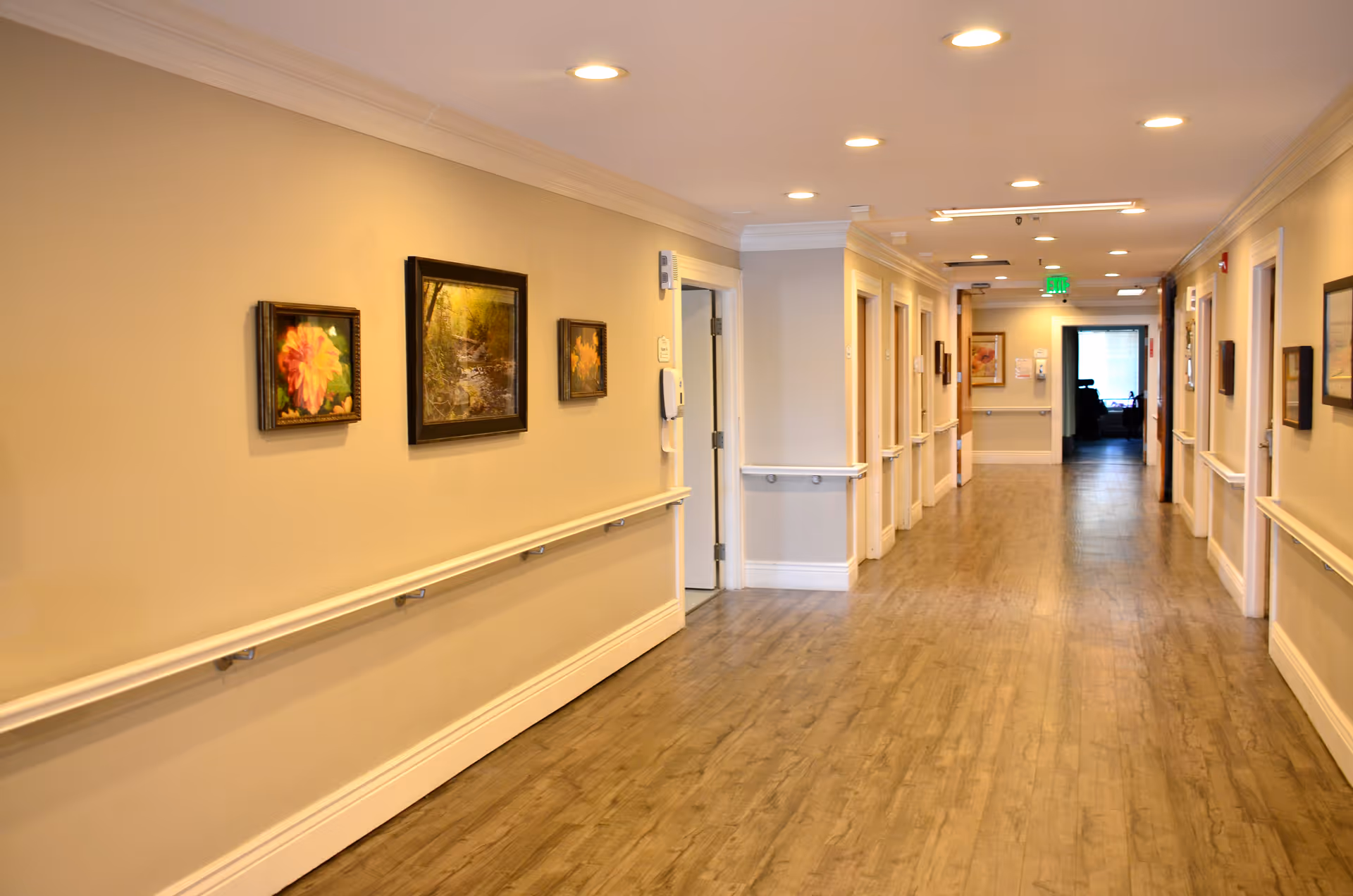 A well-lit hallway in a senior living facility with wooden flooring, beige walls, handrails on both sides, and framed pictures hanging on the walls. Several doors line the hallway, and there is an exit sign visible at the far end.
