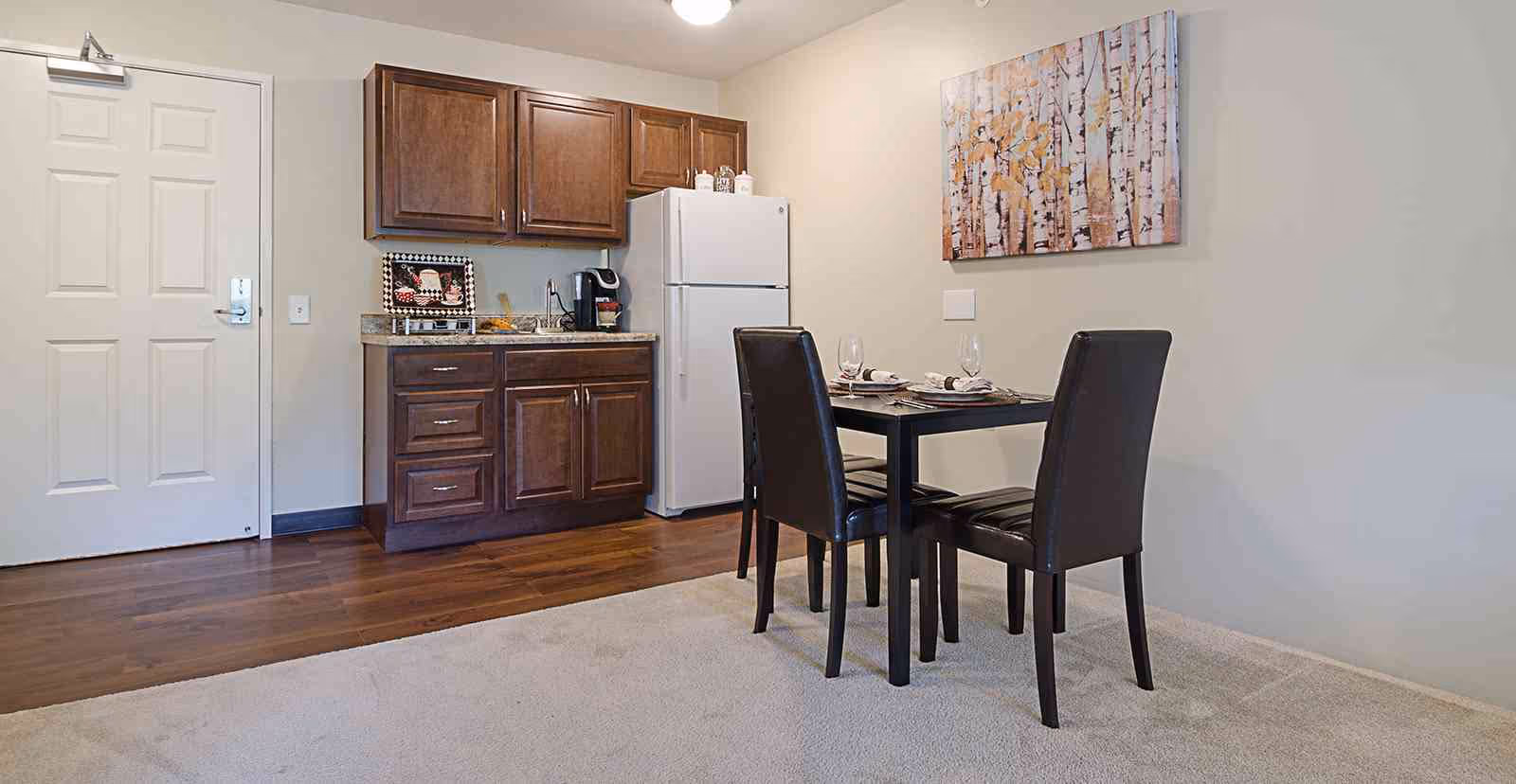 A small dining area with a black table set for two with wine glasses and napkins, adjacent to a kitchenette with wooden cabinets, a white refrigerator, a coffee maker, and a decorative tray. The room has a wooden floor near the kitchenette and carpet near the dining area, with a painting of birch trees on the wall.