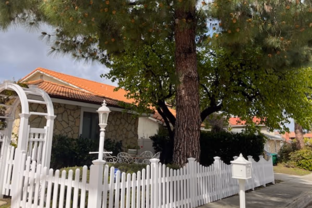 A charming outdoor scene featuring a white picket fence with a gate and a white mailbox in front of a stone house with a red tile roof. There is a large tree providing shade and white outdoor furniture visible behind the fence.