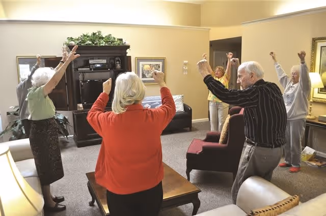 A group of elderly people standing in a living room with their arms raised, participating in a group exercise or stretching activity. The room has comfortable seating, a wooden entertainment center, framed artwork on the walls, and warm lighting.