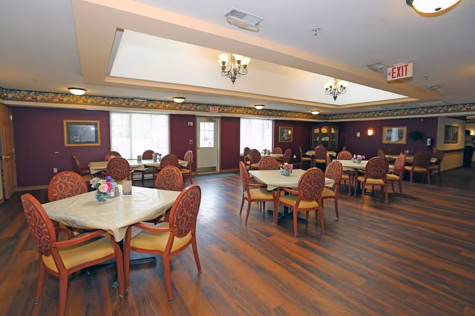 A spacious dining room with multiple tables covered with beige tablecloths and surrounded by red cushioned chairs with patterned backs. Each table has a small floral centerpiece. The room has wood flooring, purple walls with floral border trim near the ceiling, and several framed pictures on the walls. Ceiling lights and chandeliers provide illumination, and there is a door and windows along one wall.