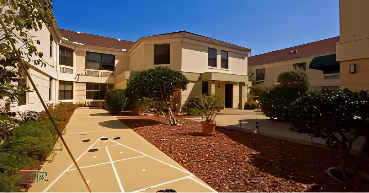 Outdoor courtyard area of a senior living facility with a shuffleboard court, surrounded by beige buildings with multiple windows, trimmed bushes, and a clear blue sky.