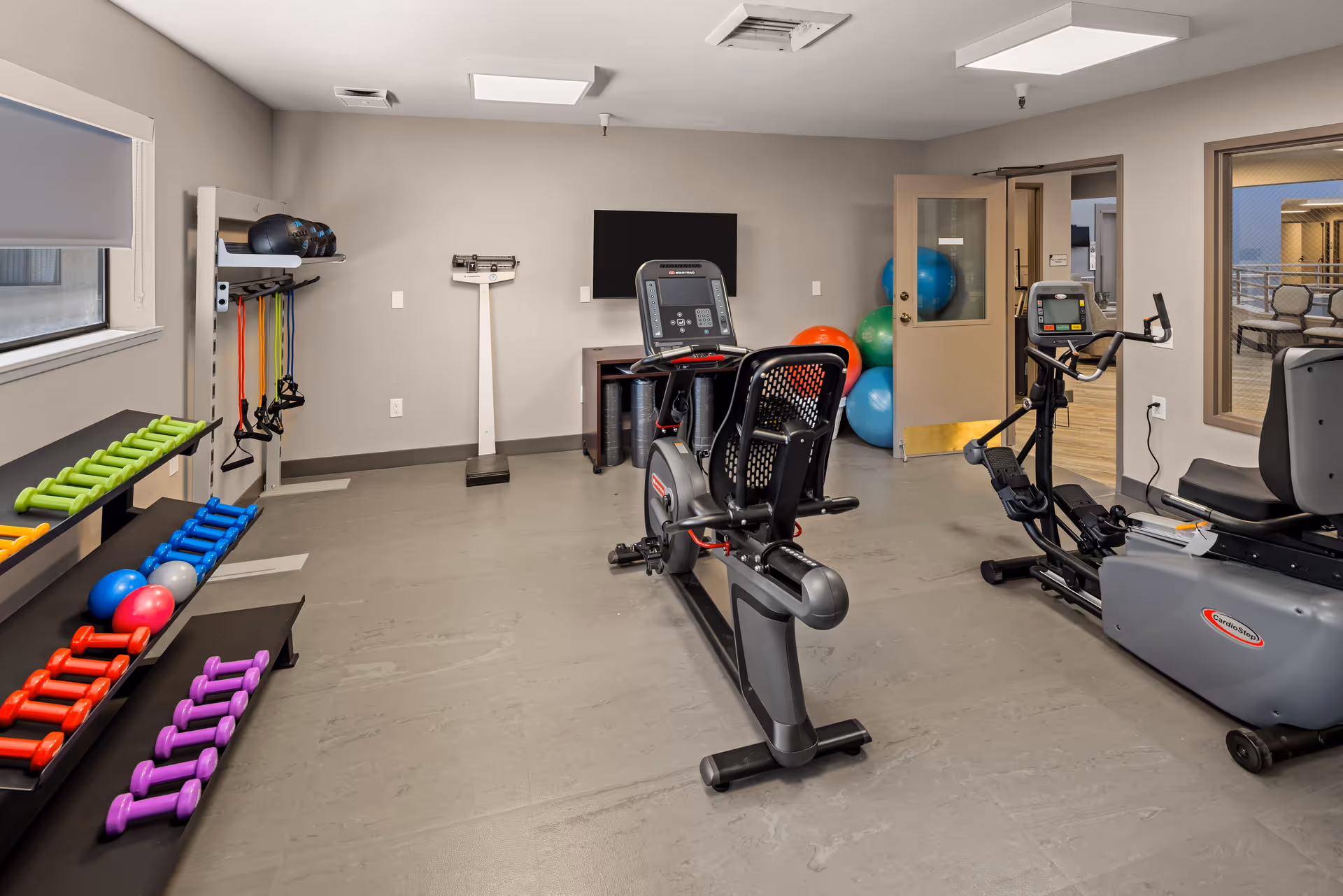 A senior living facility exercise room with various fitness equipment including colorful dumbbells on racks, resistance bands hanging on the wall, exercise balls in the corner, a stationary recumbent bike, and an elliptical machine. There is a scale against the far wall and a TV mounted above a small cabinet. The room has gray walls, a window with a blind, and a door leading to an adjacent area.