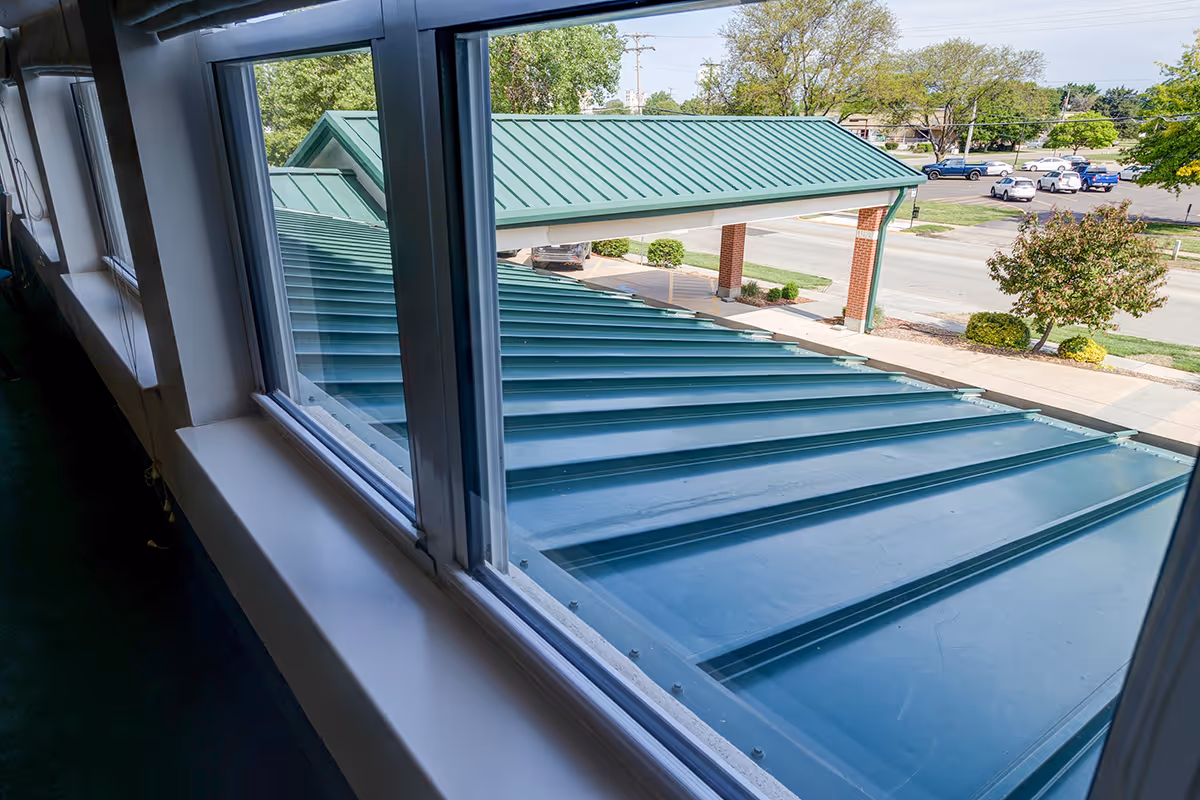 View through interior windows overlooking a green metal entrance canopy and roof with parking and landscaping beyond.