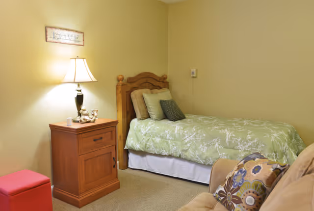 A cozy bedroom with a single wooden bed covered with a green floral bedspread and several pillows. Next to the bed is a wooden nightstand with a lamp and small decorative items. A beige armchair with a colorful floral cushion is partially visible in the foreground. The walls are painted a soft yellow color.