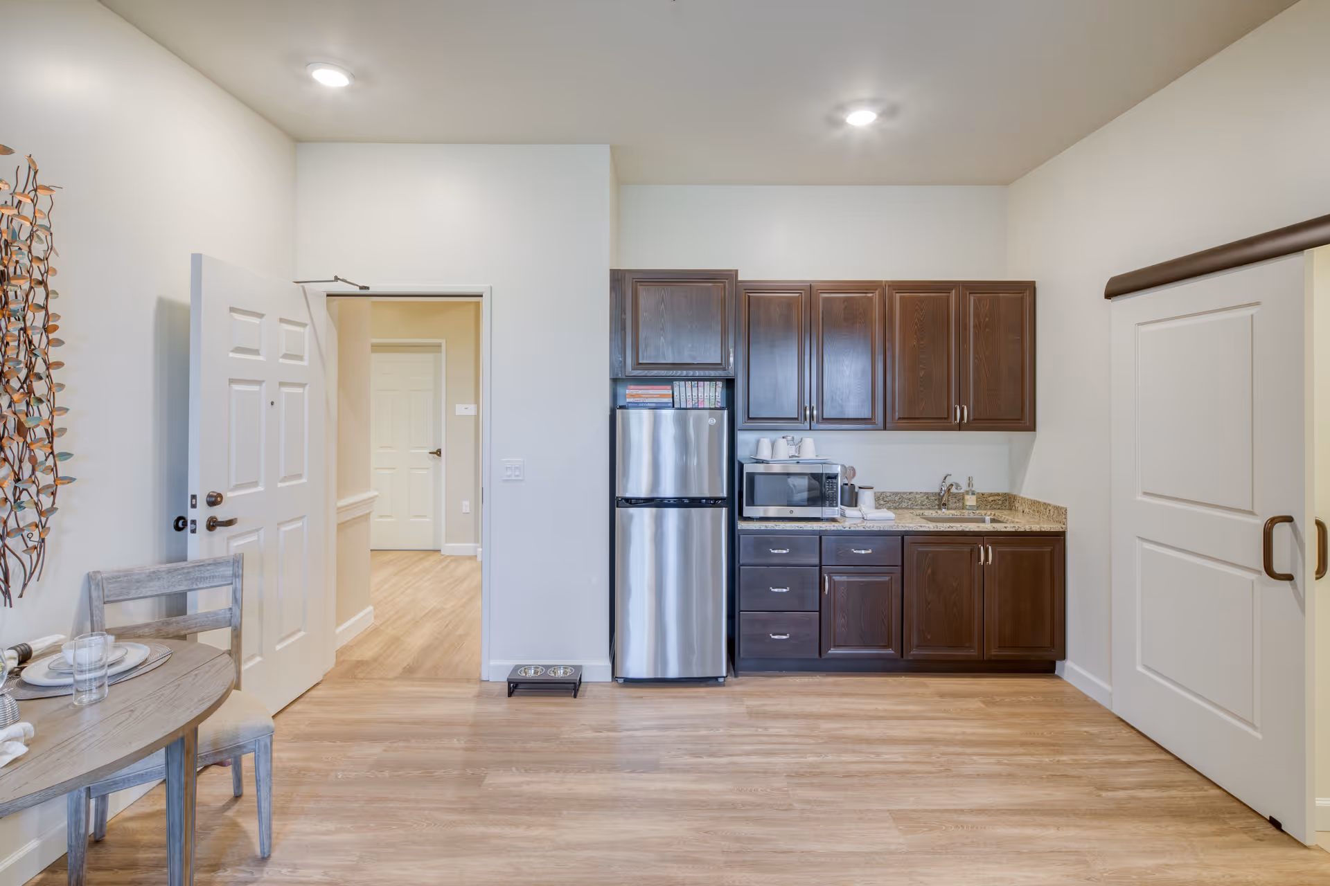 A small kitchen area with dark wooden cabinets, a stainless steel refrigerator, a microwave, and a granite countertop with a sink. To the left, there is a round wooden dining table set with plates and glasses, and a chair. The room has light-colored walls and wood flooring. A white door is open, showing a hallway with another door visible. On the right side, there is a sliding door with dark handles.