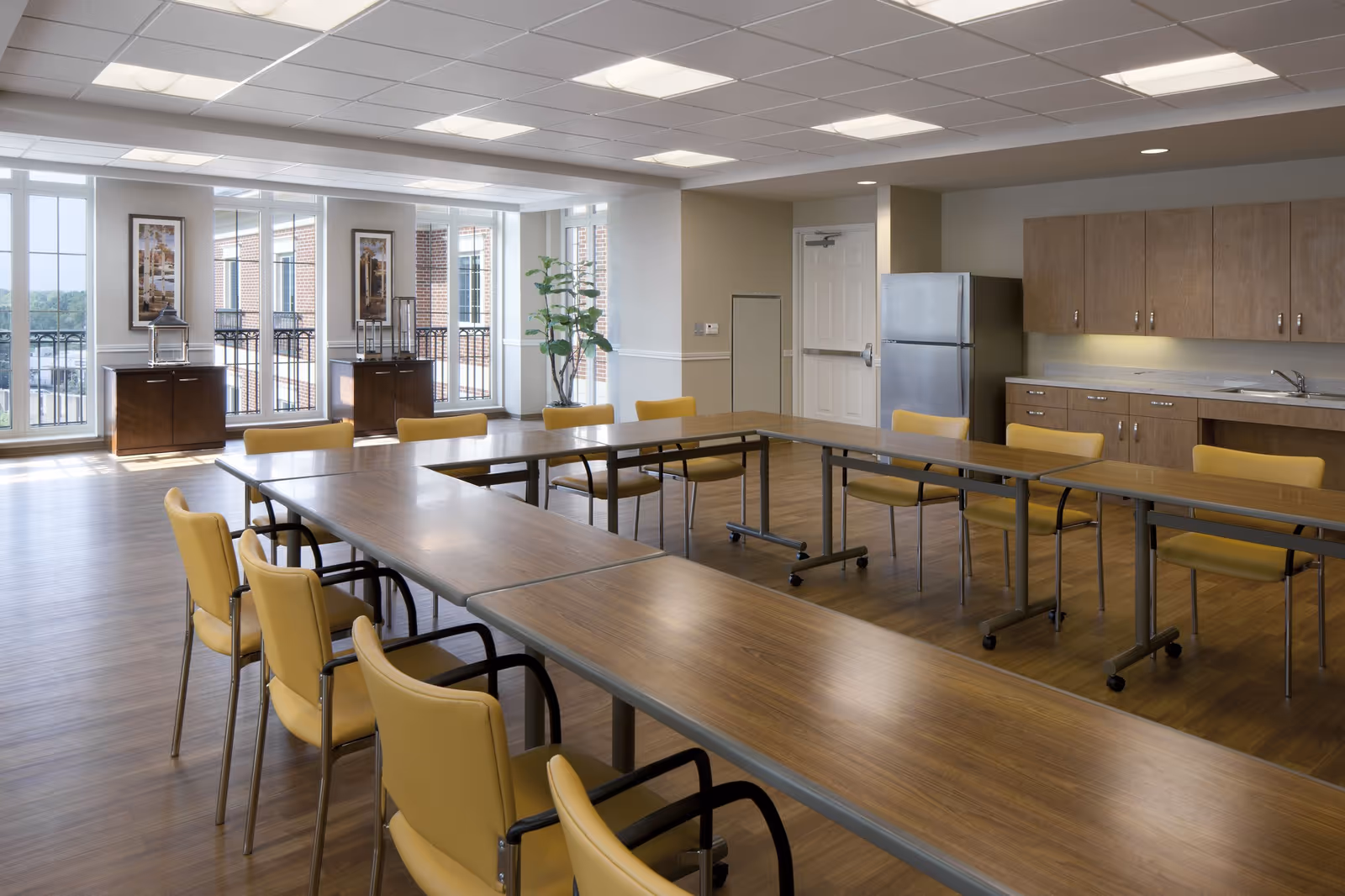 Bright multipurpose dining/meeting room with long tables, yellow chairs, a kitchenette and large windows letting in natural light.