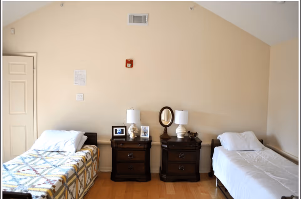 A simple bedroom with two single beds separated by two dark wooden nightstands. Each nightstand has a white lamp, and one has a small oval mirror and framed photos. The walls are beige, and the floor is wooden.