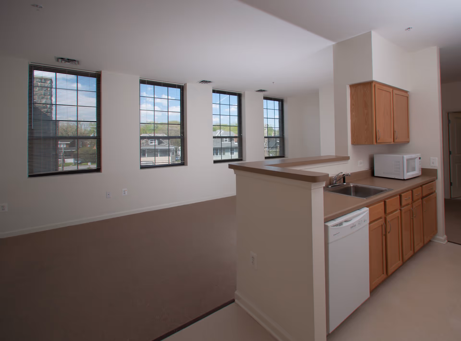 Interior view of a senior living facility unit showing a small kitchen area with wooden cabinets, a sink, a dishwasher, and a microwave on the countertop. Adjacent to the kitchen is an open living space with three large windows letting in natural light and providing a view of the outside.