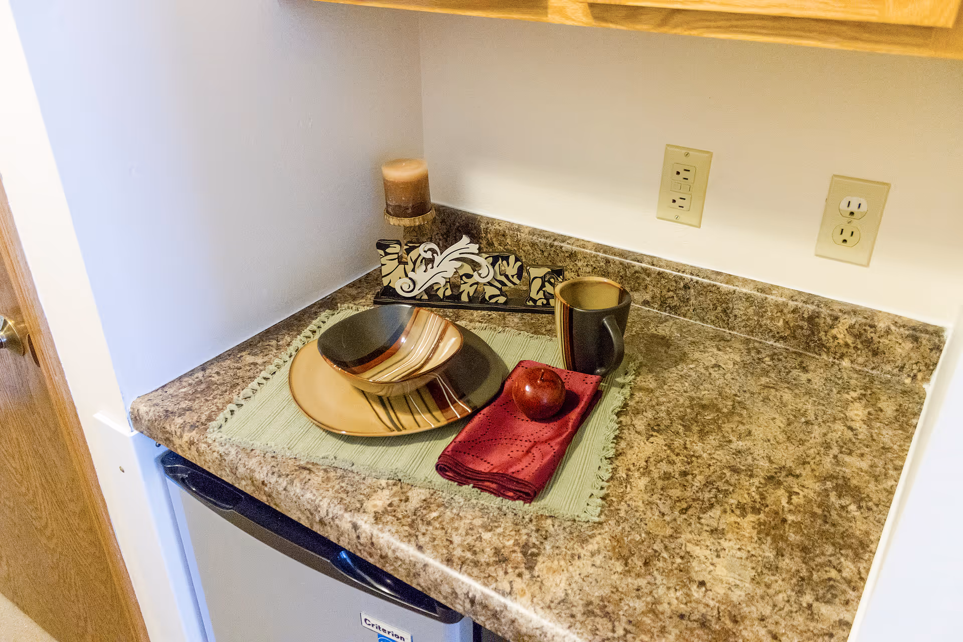 Countertop with a decorative place setting, mug and apple on a small kitchen counter above a mini fridge.