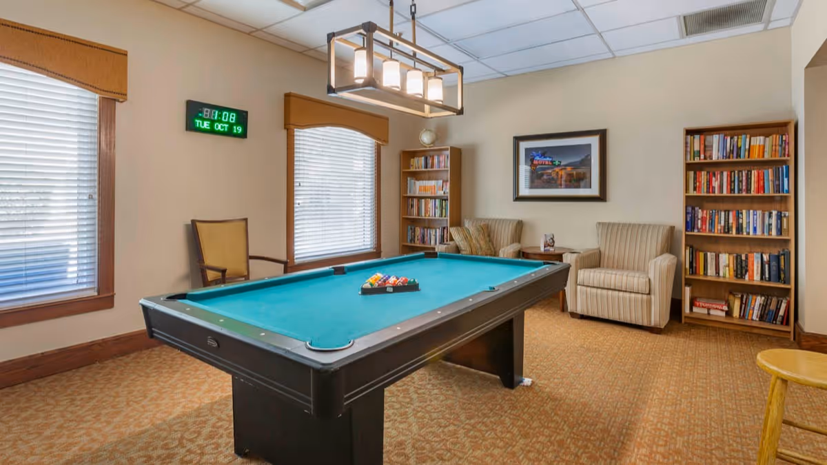 A cozy indoor room with a pool table in the center, two armchairs, two bookshelves filled with books, a framed picture on the wall, two windows with blinds and valances, and a digital clock showing the time and date.