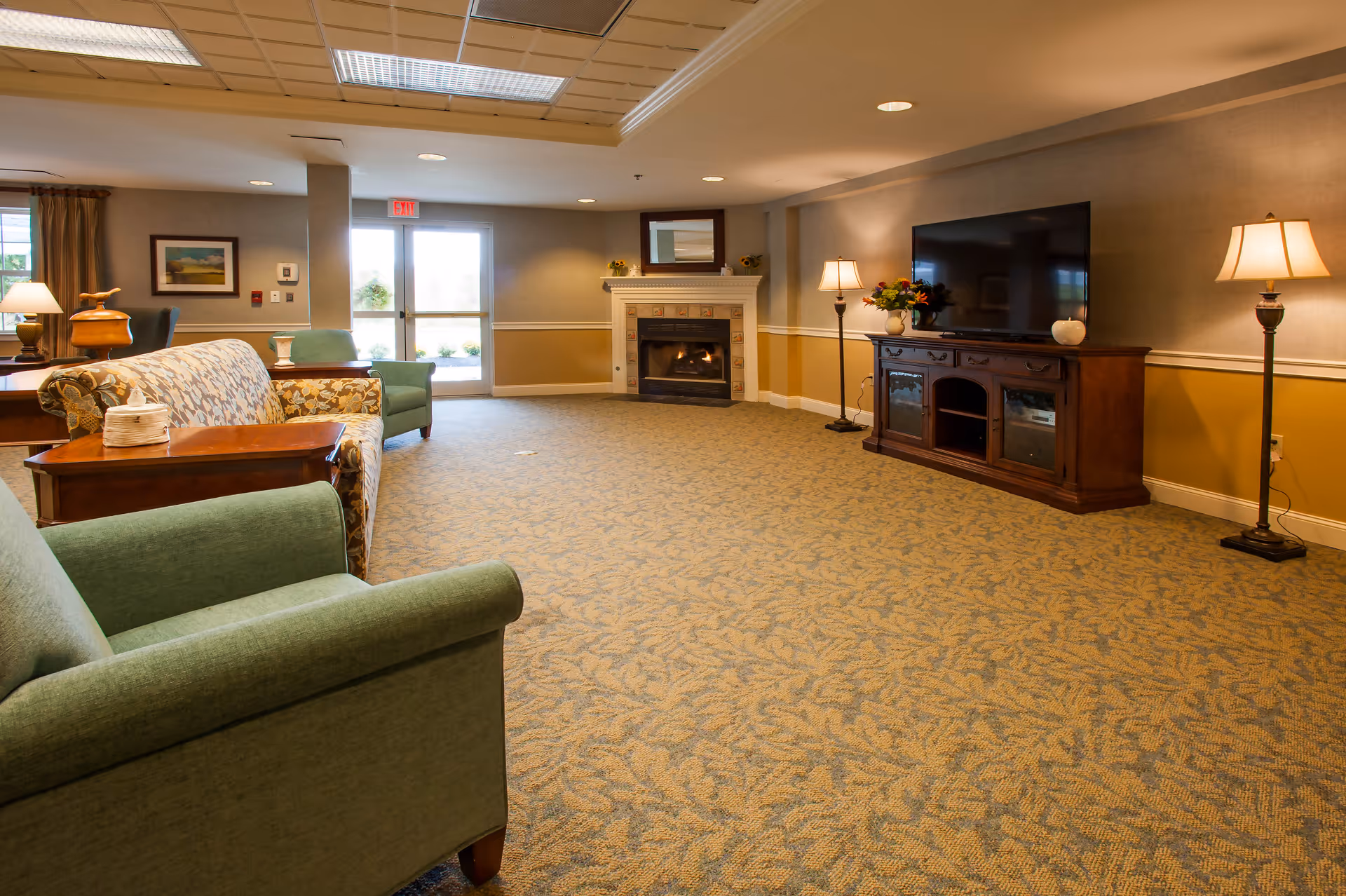 A spacious senior living common area with patterned carpet, a fireplace, a large flat-screen TV on a wooden cabinet, two floor lamps, and several upholstered chairs and sofas arranged around the room. There is a glass door leading outside and framed artwork on the walls.