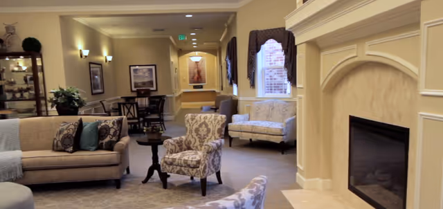 A cozy living room area in a senior living facility featuring a beige sofa with patterned cushions, an armchair with floral upholstery, a small round wooden table, a fireplace with a light-colored mantel, and a window with dark curtains. The room has soft lighting, framed artwork on the walls, and a carpeted floor.