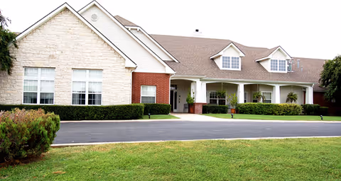 Exterior view of a single-story senior living facility building with a combination of stone and brick facade, multiple windows, a covered entrance with white columns, and a well-maintained lawn and shrubs in front.