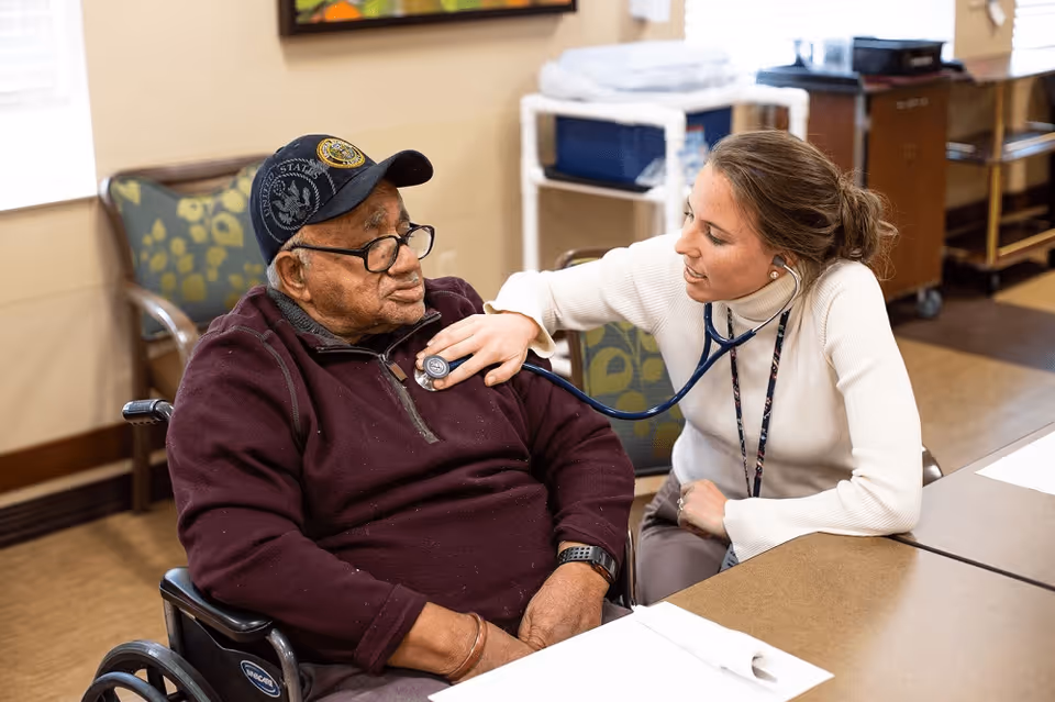 A healthcare professional uses a stethoscope to listen to the chest of an elderly man sitting in a wheelchair inside a room with chairs and tables.