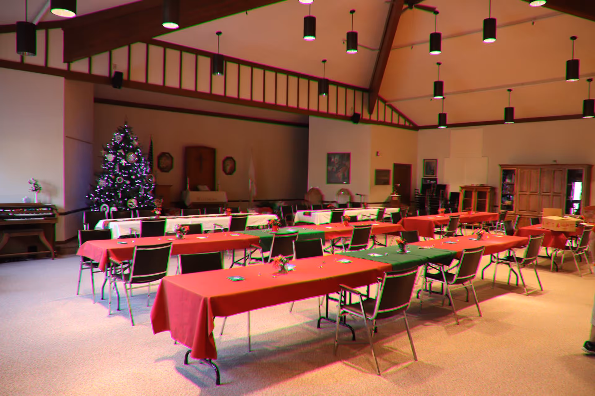 A large room set up with multiple long tables covered with red and green tablecloths, each surrounded by chairs. There is a decorated Christmas tree in the corner near a small altar with religious symbols on the wall behind it. The room has high ceilings with hanging lights and wooden beams.
