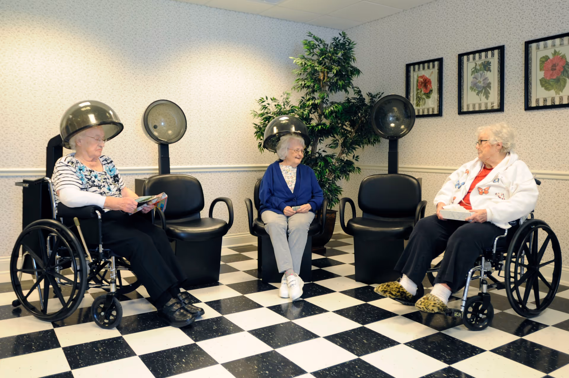 Three elderly women sitting under salon hair dryers in a room with black-and-white checkered floor and framed floral art.