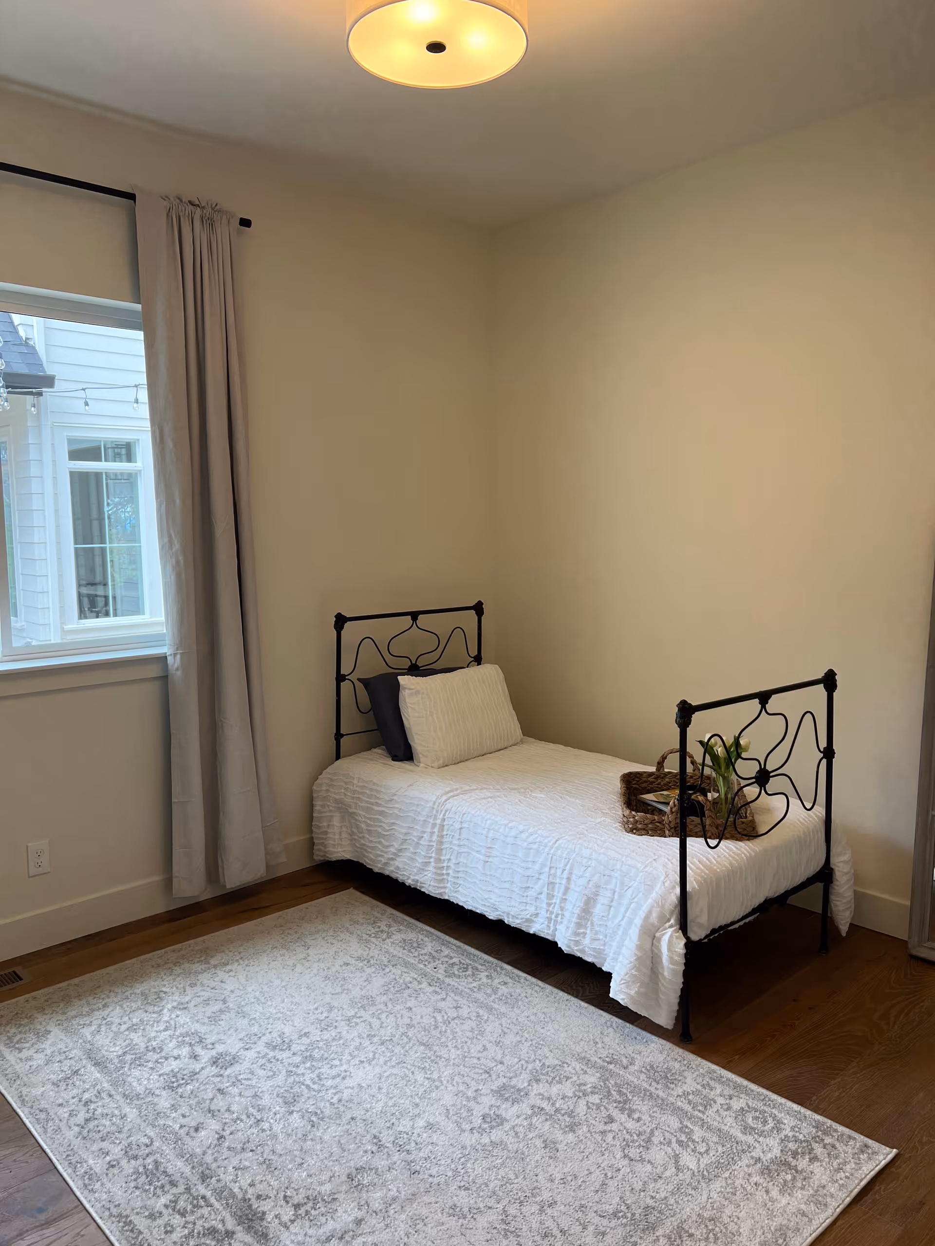 Simple bedroom with a single metal-framed twin bed, white bedding, a window with curtains, and a patterned area rug on hardwood floor.