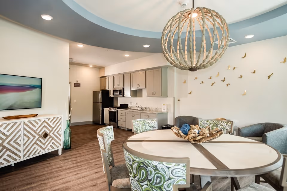 Bright communal dining area with a round table and patterned chairs, a kitchenette in the background, a large decorative chandelier and wall bird accents.
