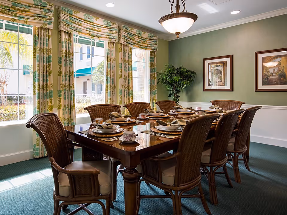 A sunlit dining room with a long wooden table set for a meal, wicker chairs, floral curtains, and framed artwork.
