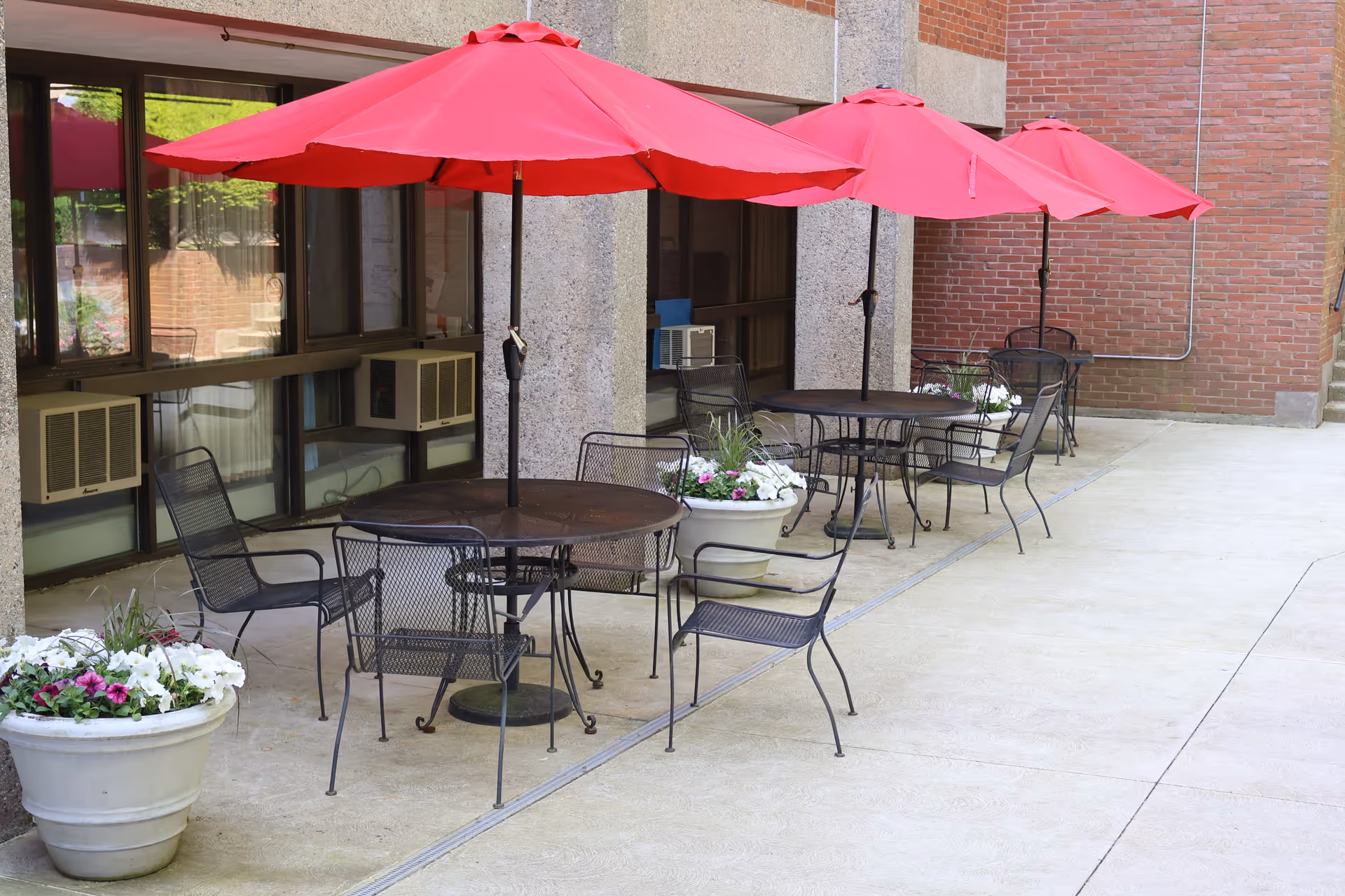 Outdoor patio area with three round metal tables, each shaded by a red umbrella. Each table is surrounded by black metal chairs. There are large white planters with flowers placed near the tables. The patio is adjacent to a building with large windows and brick walls.