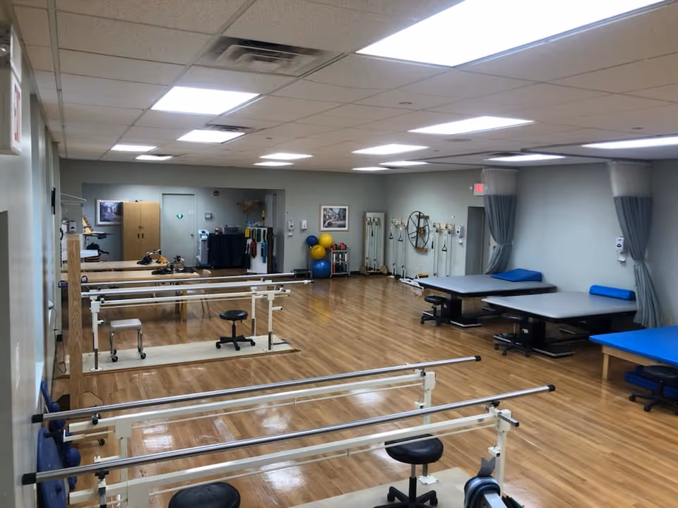 A spacious rehabilitation room with wooden floors, parallel bars for walking exercises, stools, therapy tables with blue cushions, exercise balls, and various physical therapy equipment along the walls. The room is well-lit with ceiling lights and has a calm, clinical atmosphere.