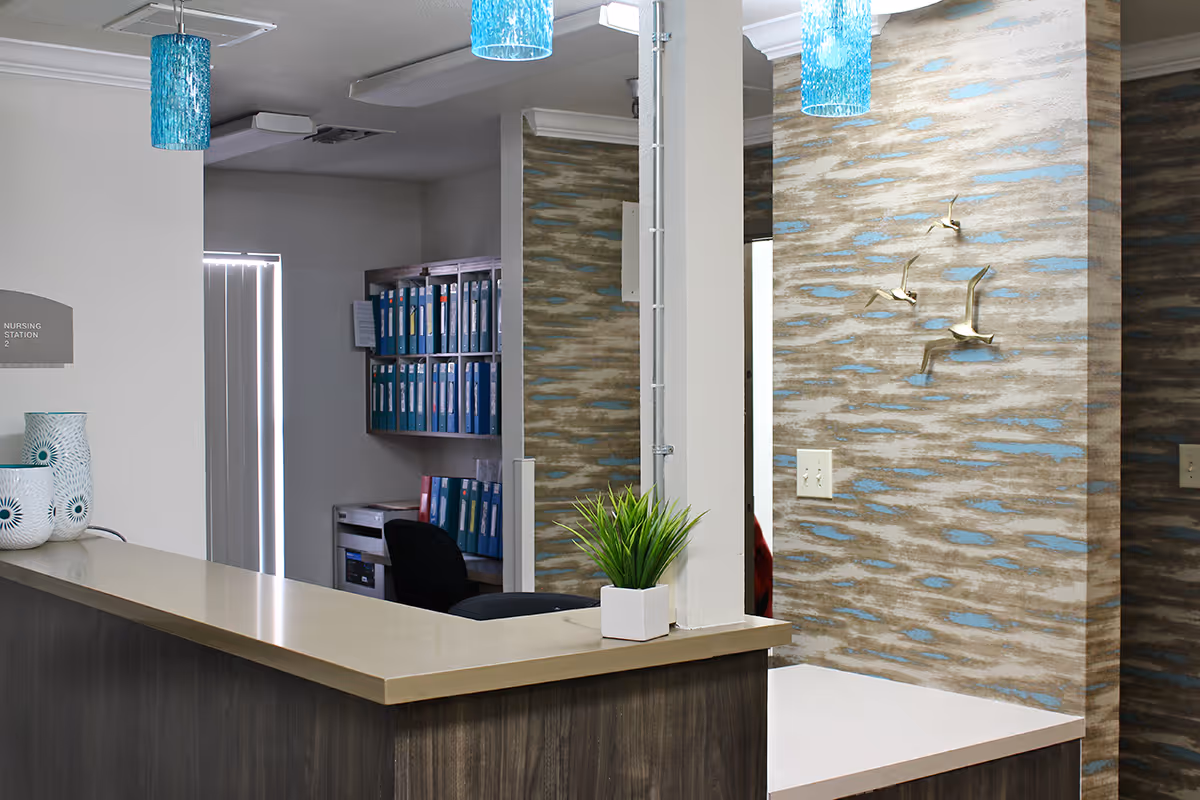 Reception area with a wooden desk and beige countertop, decorated with a small green plant in a white pot and two white vases with blue patterns. Behind the desk is a wall with blue and beige textured wallpaper and three metallic bird sculptures mounted on it. Blue pendant lights hang from the ceiling. In the background, there is a doorway leading to an office area with shelves filled with blue binders and a black office chair.