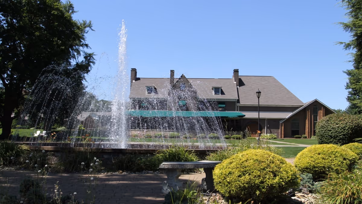 A large brick building with a steep roof and multiple chimneys is seen behind a water fountain spraying water high into the air. The foreground features a stone bench surrounded by well-maintained bushes, plants, and a paved walkway under a clear blue sky.