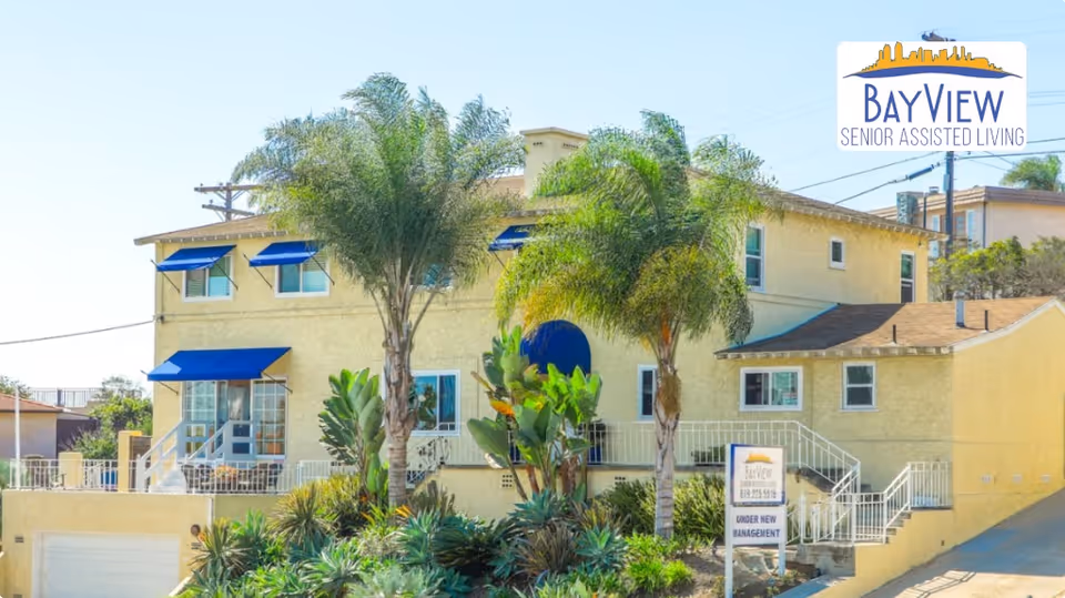 Exterior view of a two-story yellow building with blue window awnings and palm trees in front, identified as BayView Senior Assisted Living. There is a sign in front of the building indicating it is under new management.