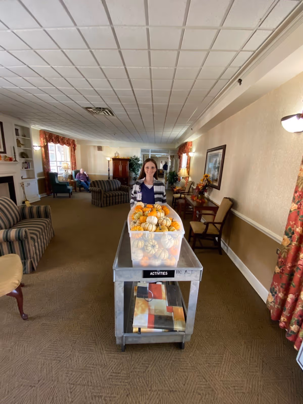 A woman standing behind a metal cart labeled 'ACTIVITIES' filled with small decorative pumpkins in a cozy living room area. The room has striped armchairs, a fireplace, lamps, framed artwork, and floral curtains. Another person is seated in the background near a window.