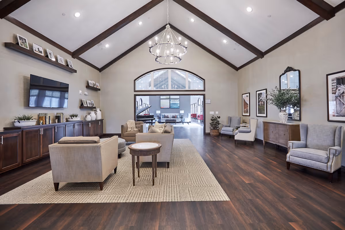 A spacious and well-lit living room with high vaulted ceilings and dark wooden beams. The room features a large area rug with beige armchairs and a small round wooden table in the center. On the left wall, there is a mounted flat-screen TV above dark wooden cabinets decorated with plants and framed photos. The right wall has framed artwork, a large mirror, and additional armchairs. Through a large arched doorway, another room with a grand piano and more seating is visible.