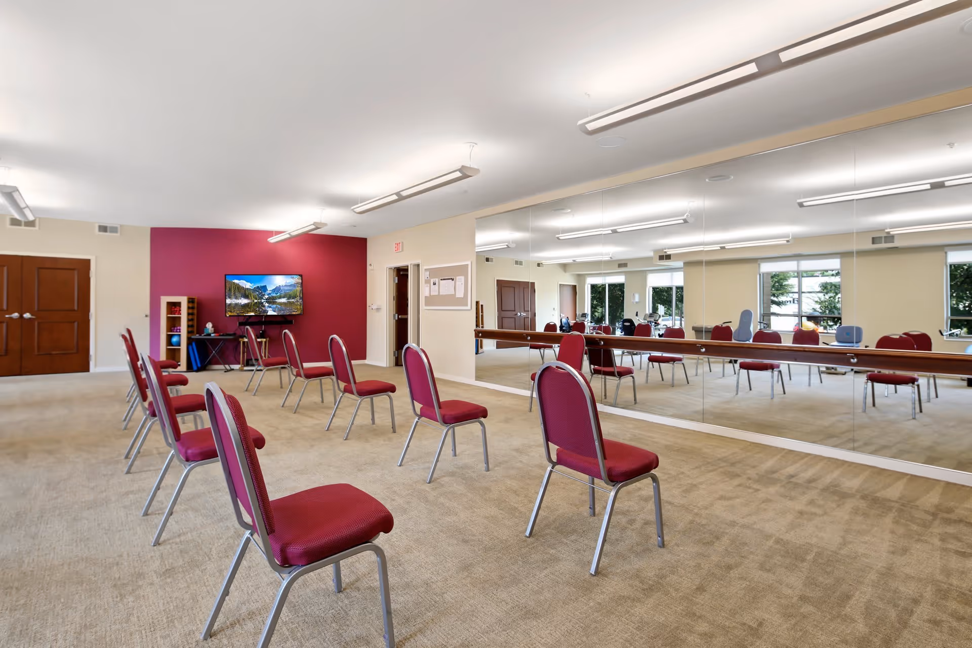 A spacious room with beige carpet and cream-colored walls, featuring a large wall mirror on one side. Several red cushioned chairs with metal legs are arranged in rows facing a wall-mounted TV on a burgundy accent wall. The room is well-lit with ceiling lights and has windows letting in natural light.