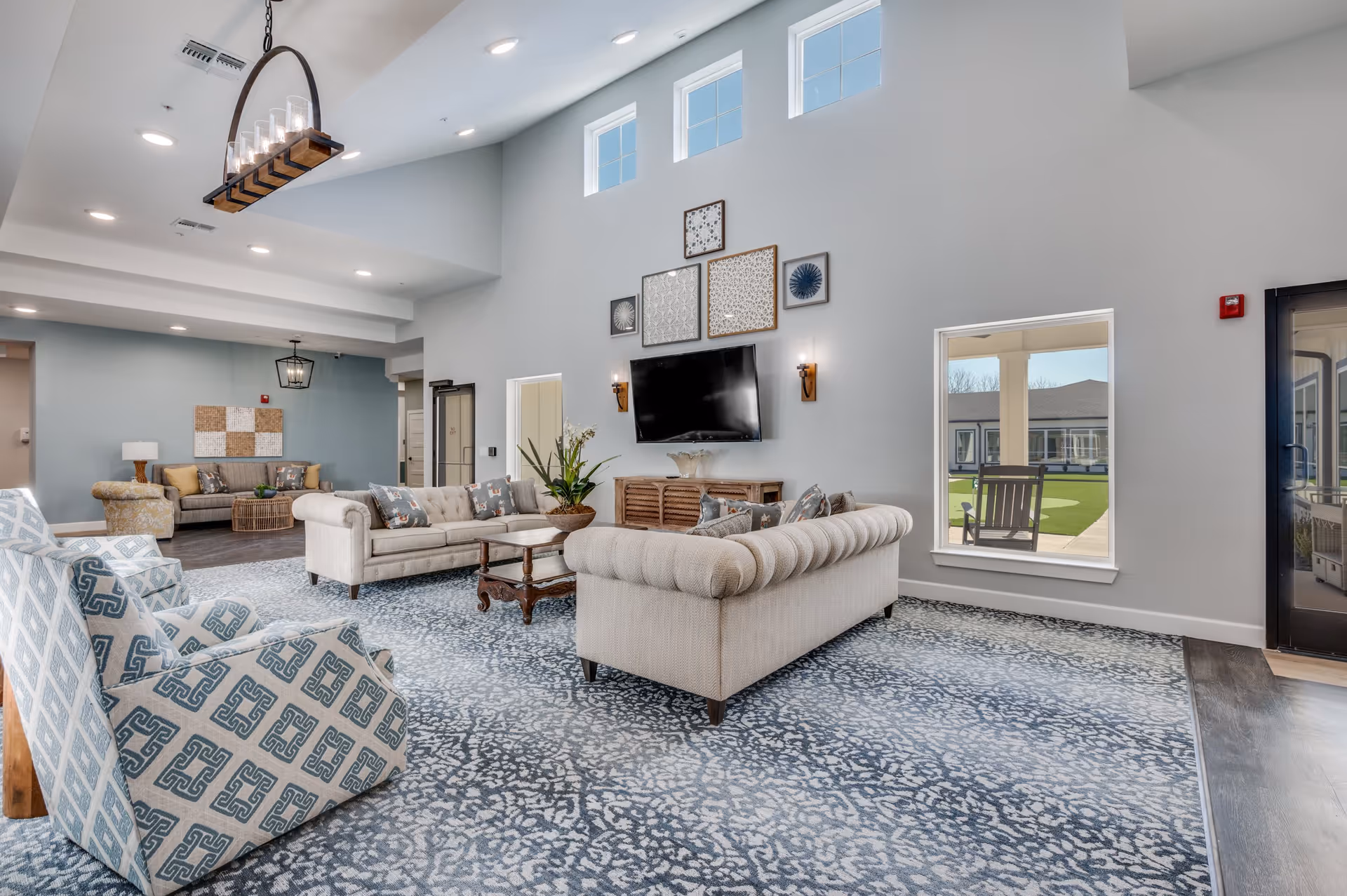 A spacious and well-lit living room area with high ceilings and multiple windows near the ceiling. The room features two beige tufted sofas arranged around a wooden coffee table, two patterned armchairs in the foreground, and a TV mounted on the wall above a wooden console. Decorative wall art is displayed above the TV. A large patterned area rug covers the floor, and there is a view through a window and glass door to an outdoor patio with chairs and green lawn.