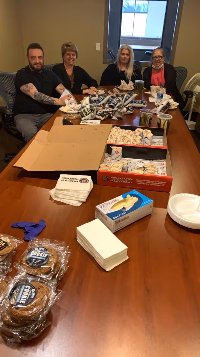 Four people sitting around a wooden conference table with various snacks, including cookies, sandwiches, and drinks, spread out on the table in a meeting room with a window in the background.